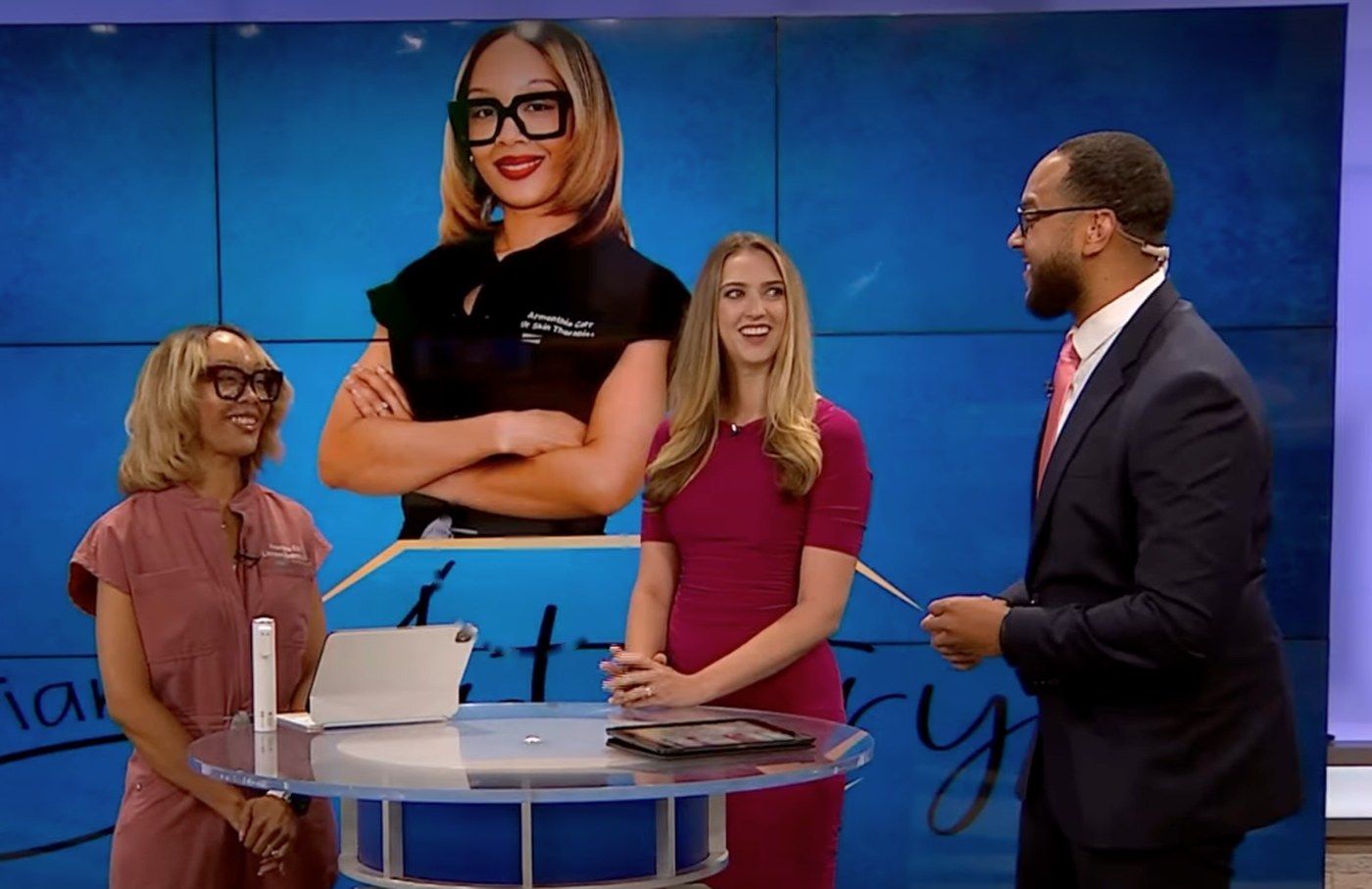 Armenthia Carr on a television news set with two anchors, her portrait and the IAMA Artistry logo on the screen behind them