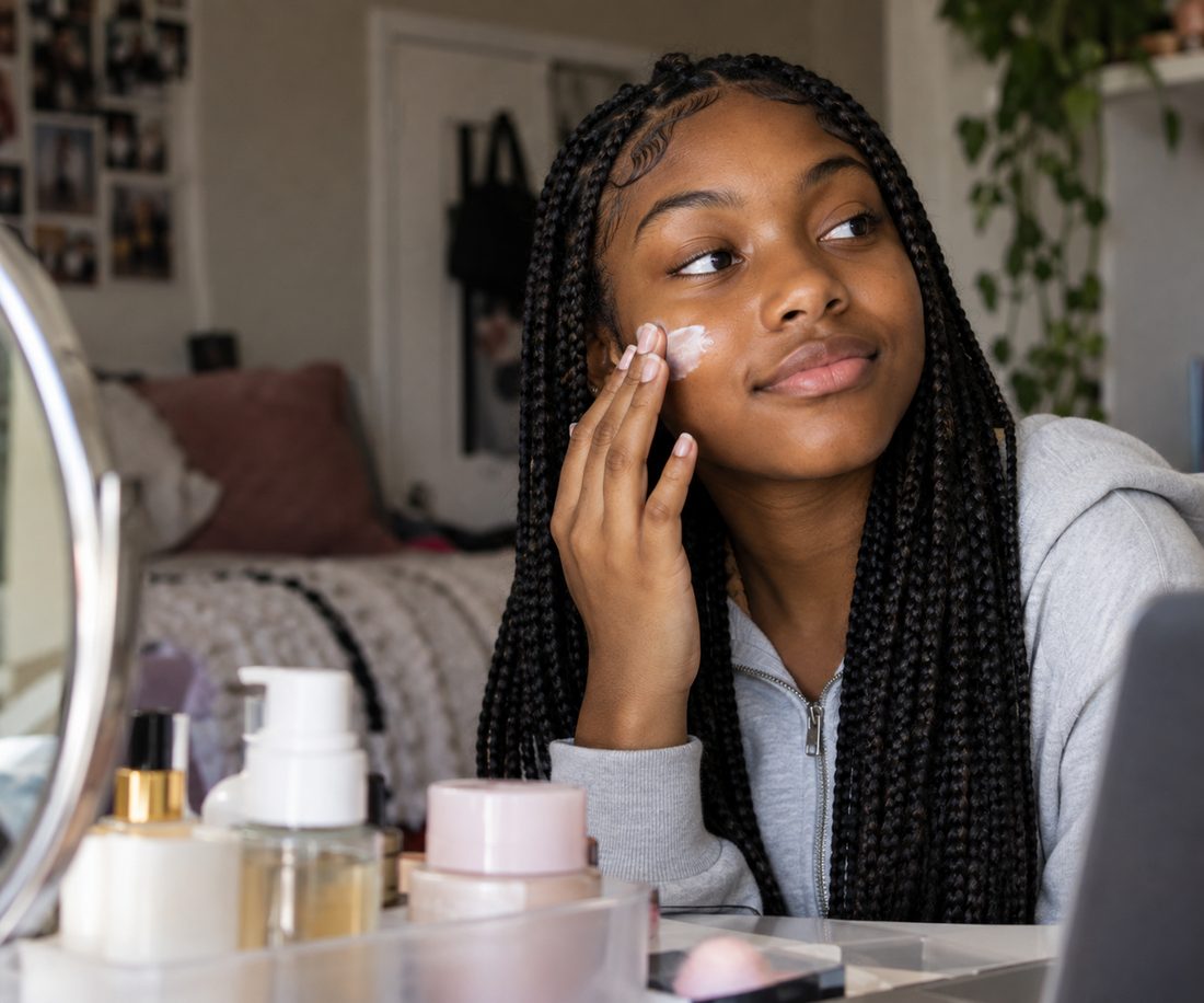 A teenager applies moisturizer at a vanity crowded with skincare bottles and jars.